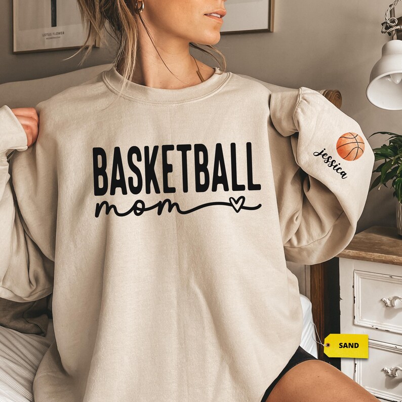 a woman sitting on a bed wearing a basketball mom sweatshirt