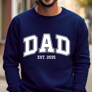 May include: Navy blue sweatshirt with the word "DAD" in large, white, block letters. Below the word "DAD" is the text "EST. 2025" in white. The sweatshirt is a solid color and has a crew neck.