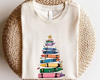 Könnte beinhalten: Cremefarbenes T-Shirt mit einem Weihnachtsbaum-Design aus bunten Buntstiften. Die Buntstifte sind zu einem Baum geformt, verziert mit Lichterketten und einem goldenen Stern. Das Shirt ist aus weichem Material.