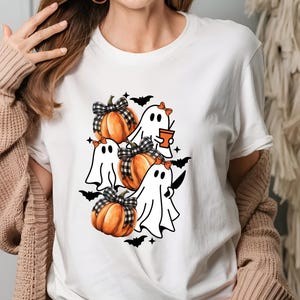 May include: White t-shirt featuring a Halloween-themed design. The graphic includes orange pumpkins with black and white plaid bows, cartoon ghosts, and black bats. One ghost holds a cup, another a knife. A fun, festive design.