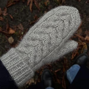 Puede incluir: Un mitón tejido de color gris claro con un diseño de punto de cable. El mitón está hecho de un hilo suave y texturizado. El fondo muestra hojas caídas y una acera pavimentada.
