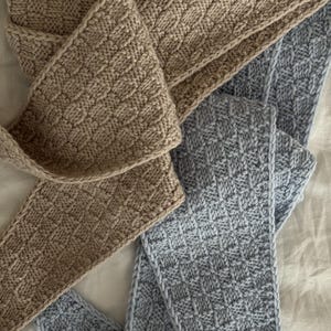 Può includere: Primo piano di due sciarpe lavorate a maglia. Una è di colore beige chiaro, l'altra è azzurra. Entrambe le sciarpe hanno un motivo strutturato e intrecciato. Le sciarpe sono realizzate in un materiale morbido e caldo.