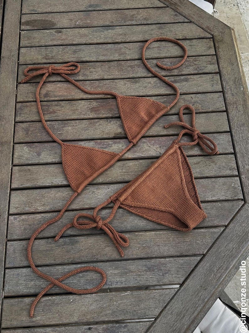 May include: A brown knitted bikini set, featuring a triangle top and a low-rise bottom. The bikini has tie closures and is displayed on a wooden surface. The overall design is simple and minimalist.