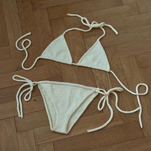 May include: Cream-colored knitted bikini set. The triangle top and low-rise bottoms feature tie closures. The swimwear is made of a textured, soft material, perfect for a beach or poolside look.