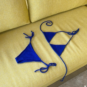 May include: A blue knitted bikini top and bottom set. The triangle top and bottom are connected with thin ties. The bikini is displayed on a yellow sofa.