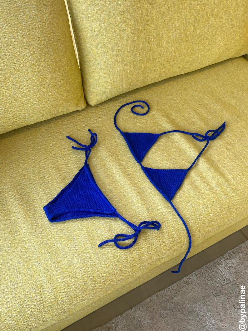 May include: A blue knitted bikini top and bottom set. The triangle top and bottom are connected with thin ties. The bikini is displayed on a yellow sofa.