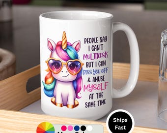 May include: White ceramic mug featuring a cartoon unicorn with heart-shaped sunglasses and a rainbow mane. The mug has the text "PEOPLE SAY I CAN'T MULTITASK BUT I CAN PISS YOU OFF & AMUSE MYSELF AT THE SAME TIME".