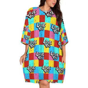 May include: A loose-fitting dress featuring a vibrant checkered pattern in yellow, red, and turquoise squares. The fabric is printed with the repeated black and white text "LOVE YOU" in a bold, stylized font across the entire garment surface.