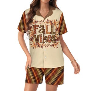 May include: A short-sleeve button-down shirt and matching shorts set in a cream color. The shirt features a brown collar and a graphic print of autumn leaves surrounding the text FALL VIBES in a plaid pattern. The sleeves and shorts are plaid.