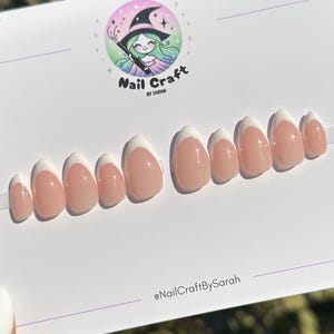 May include: A set of French tip press-on nails with a nude base and white tips. The nails are arranged on a white surface with the text "Nail Craft by Sarah" and a cartoon witch logo.