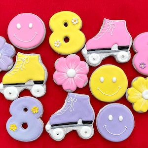 Puede incluir: Una colección de galletas decoradas sobre un fondo rojo. Las galletas tienen varias formas, incluyendo patines, caras sonrientes, flores y el número ocho. Las galletas están decoradas con glaseado rosa, amarillo, morado y blanco.