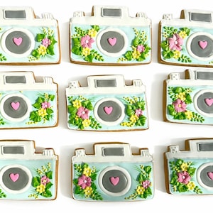 May include: Nine camera-shaped cookies with light blue bodies, gray lenses, and white tops. Each cookie is decorated with pink, yellow, and green floral designs and a pink heart in the center of the lens. The cookies are arranged on a white surface.