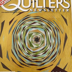 May include: The cover of Quilters Newsletter magazine, June/July 2013 edition. The cover features a geometric quilt design with concentric circles in shades of gold, green, blue, and brown. The magazine title and issue details are visible.