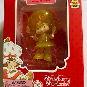 May include: A Lemon Meringue figure from The World of Strawberry Shortcake, packaged in a red box. The figure is yellow and holding a mirror. The box has the TLS Toy logo and a warning about small parts.