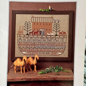 May include: A framed cross-stitch sampler titled "Noah's Ark Sampler" with a brown wooden frame. The sampler depicts Noah's Ark, animals, and text. Two wooden camel figurines and greenery are in the foreground.