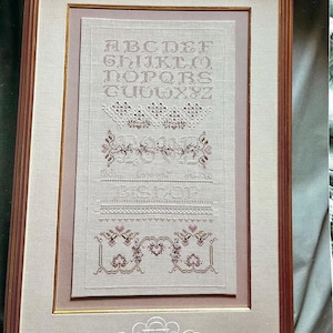 May include: A framed cross-stitch sampler, titled "The Love Sampler." The design includes an alphabet, decorative borders, and the name "BISHOP" stitched in brown thread on a light beige fabric. The frame is brown with a cream-colored mount.