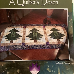 Op de afbeelding: Een quiltpatroonboek getiteld "Table Runners: A Quilter's Dozen" van Brenda Henning. De omslag van het boek toont een foto van een tafelkleed met een paars, bruin en groen patchwork-ontwerp met drie gestileerde dennenbomen.