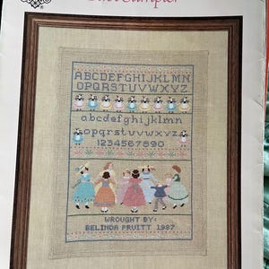 May include: A framed cross-stitch sampler titled "A Girl's First Sampler." The design features the alphabet, numbers, and figures of children in dresses. The sampler is in a wooden frame and includes the text "Wrought by: Belinda Pruitt 1987."