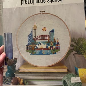May include: A cross-stitch embroidery hoop featuring a colorful Sydney, Australia cityscape. The design includes the Sydney Opera House, Harbour Bridge, and other landmarks. The text "pretty little sydney" is at the top.