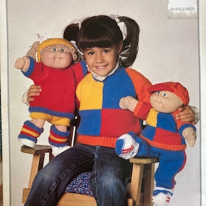 Könnte beinhalten: Ein Mädchen in blauen Jeans sitzt auf einem Holzstuhl und hält zwei Cabbage Patch Kids Puppen. Die Puppen tragen bunte gestrickte Outfits. Das Mädchen trägt einen bunten Strickpullover. Das Bild stammt aus einem Vintage-Strickmusterbuch.
