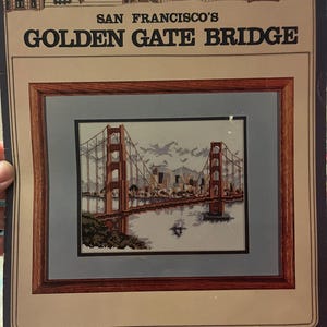May include: A cross-stitch kit featuring a framed image of the Golden Gate Bridge in San Francisco. The kit includes the text "SAN FRANCISCO'S GOLDEN GATE BRIDGE" and "COUNTED CROSS-STITCH by DEBBIE PATRICK". The bridge is depicted in shades of brown and gray.