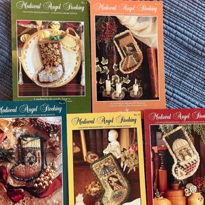 May include: Five books titled "Medieval Angel Stocking" are shown, each with a different coloured cover. The covers feature images of cross-stitch Christmas stockings. The books are in shades of green, orange, yellow, and red, with detailed designs.