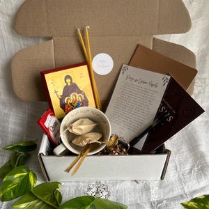 Pode incluir: Uma caixa de cartão castanho cheia de uma variedade de itens, incluindo um ícone religioso emoldurado, duas velas amarelas, um pequeno caderno, um cartão com o texto "Oração para Pais Urbanos", uma caneca com saquetas de chá e uma barra de chocolate. A caixa está sobre uma superfície branca.