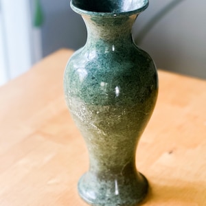 May include: A green marble vase with a flared rim and a rounded base. The vase has a smooth, polished surface and a classic, elegant shape. The color is a mix of dark and light greens.