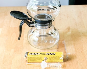 Op de afbeelding: Een vacuüm koffiezetapparaat van helder glas met een zwarte kunststof handgreep en deksel, voorzien van horizontale zwarte strepen. Daarnaast ligt een gele doos met het opschrift CORY GLASS FILTER ROD en een glazen filterstaaf.