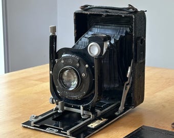 May include: A vintage black folding camera with a bellows mechanism, a circular lens assembly, and a metal frame. The camera features a top-mounted handle, a silver shutter release button, and a separate black rectangular plate component nearby.