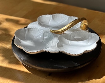 May include: A white, three-section serving dish with a gold handle and gold trim. The dish sits on a dark, round wooden base. The dish's sections are shaped like flower petals.