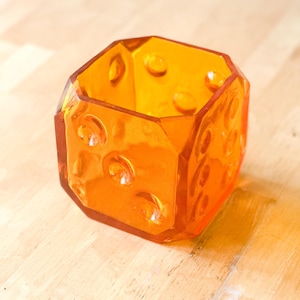 May include: An orange, geometric-shaped glass container with a faceted design. The sides feature circular, raised details. The container sits on a light-colored wooden surface. The object is empty and appears to be a decorative item.