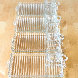 Könnte beinhalten: Vier Snack-Sets aus klarem Glas, bestehend aus einem rechteckigen Tablett mit gerippter Textur und einer passenden runden Tasse. Die Tabletts haben kleine Griffe an den Seiten und die Tassen verfügen über ein markantes Perlenhenkel-Design.