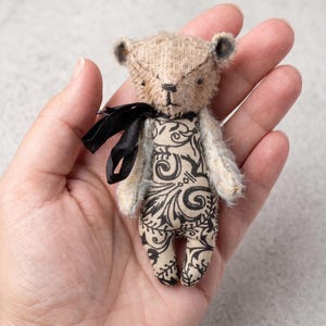 May include: A miniature teddy bear with a textured beige head and arms, featuring a black ribbon tied around its neck. The body is made of a light-coloured fabric with a detailed black floral pattern. The bear has small dark eyes and a stitched nose.