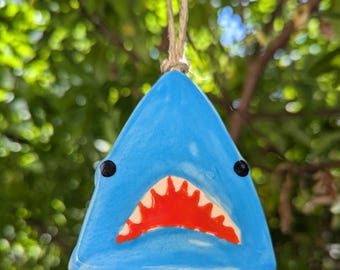 May include: A blue ceramic shark ornament with a red open mouth and black eyes. The ornament is hanging from a string.