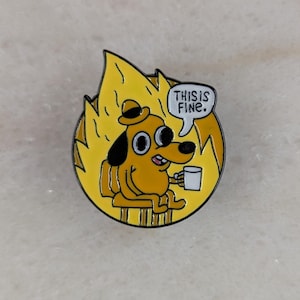 May include: A yellow enamel pin depicting a cartoon dog in a burning room, holding a coffee cup. The dog wears a hat and the speech bubble reads "THIS IS FINE."