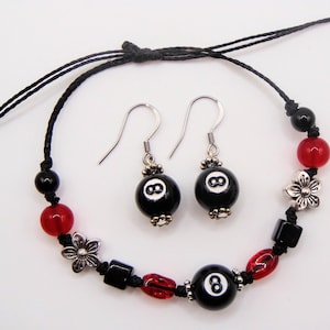 May include: A jewelry set consisting of a black cord bracelet and two matching drop earrings. The bracelet features a central black 8-ball bead, red glass beads, black square beads, and silver-toned flower charms. The earrings feature 8-ball beads.