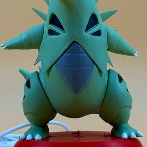May include: A green Tyranitar figure with a wide open mouth, standing on a red, black, and white Poké Ball base. The figure has sharp spikes and a detailed design. The base has the text "TOONTRONIX".