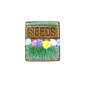 May include: A square pin featuring a brown seed packet design with the word "SEEDS" in gold lettering. The packet has a green base with colourful flowers and grass.