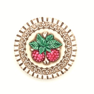 May include: A round ceramic coaster with a raised design of two red raspberries and green leaves. The coaster has a brown and white border with a decorative pattern.