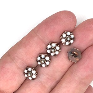 May include: A collection of antique-style buttons. Each button has a hexagonal shape with a bronze-colored metal frame. The center features a small, round pearl-like bead surrounded by white, circular accents.