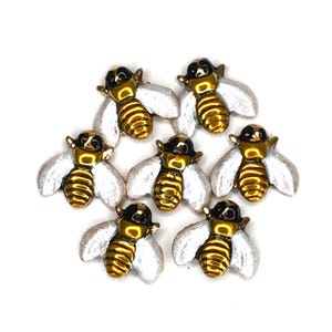 May include: Seven decorative bee-shaped embellishments. Each bee features a gold-colored body with black accents and white wings. The bees are arranged in a circular pattern against a white background, creating a visually appealing design.