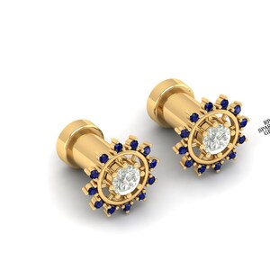 以下が含まれることがあります： 中央に透明な石があり、小さな青い石で囲まれた、金色のイヤリングのペア。 イヤリングは円筒形のポストと、太陽光のような装飾的なデザインをしています。 背景は白です。