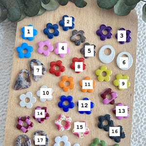 May include: A collection of colorful flower-shaped beads in various colors, including blue, pink, red, orange, and white. Some beads have a marbled effect. The beads are arranged on a wooden cutting board.