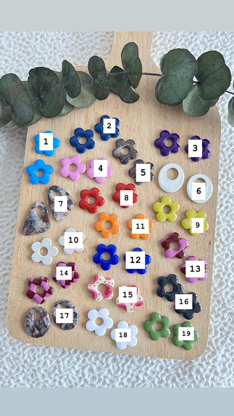 May include: A collection of colorful flower-shaped beads in various colors, including blue, pink, red, orange, and white. Some beads have a marbled effect. The beads are arranged on a wooden cutting board.