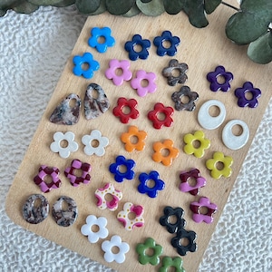 May include: A collection of colorful flower-shaped earrings and other jewelry components. The earrings are in various colors, including blue, pink, red, orange, yellow, and green. Some have patterned designs. The items are arranged on a wooden cutting board.
