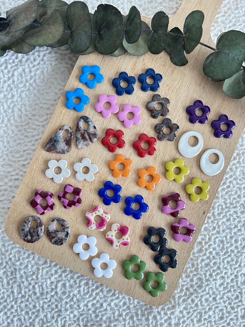 May include: A collection of colorful flower-shaped earrings and other jewelry components. The earrings are in various colors, including blue, pink, red, orange, yellow, and green. Some have patterned designs. The items are arranged on a wooden cutting board.
