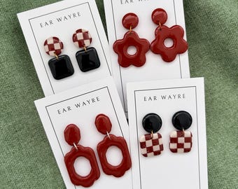 May include: Four pairs of polymer clay earrings on white display cards labeled EAR WAYRE. Designs include red and white checkered circles with black squares, solid red flower shapes, red wavy ovals, and black circles with red and white checkered squares.
