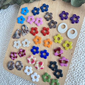 May include: A collection of colorful flower-shaped earrings in various colors, including blue, pink, red, orange, yellow, and green. Also included are marbled and patterned earrings, and white hoop earrings. The earrings are displayed on a wooden board.