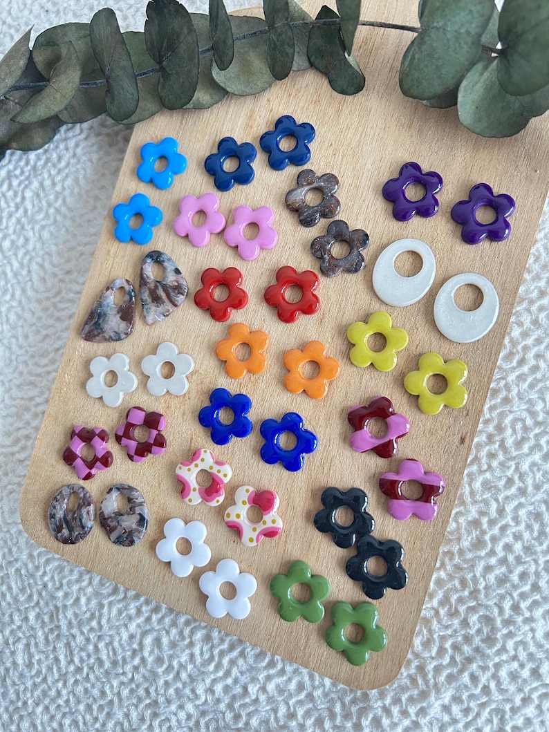 May include: A collection of colorful flower-shaped earrings in various colors, including blue, pink, red, orange, yellow, and green. Also included are marbled and patterned earrings, and white hoop earrings. The earrings are displayed on a wooden board.